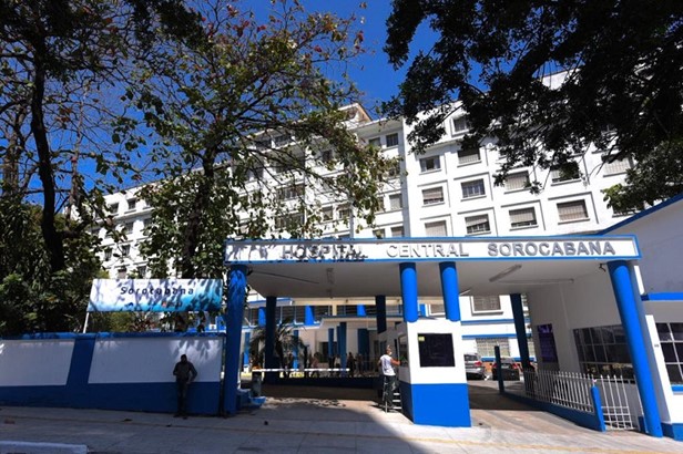 FOTO DE EDIFICAÇÃO - Um grande prédio branco com muro e detalhes em azul . Na fachada temos a sinalização Hospital Sorocabana. Há árvores na calçada e pessoas entrando  no local